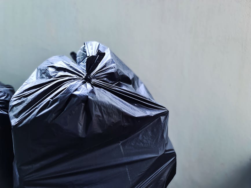 A worker wearing a red high-visibility vest is seen emptying waste into the rear compartment of a large red refuse collection vehicle on the side of a street. The waste appears to be a mix of black pl