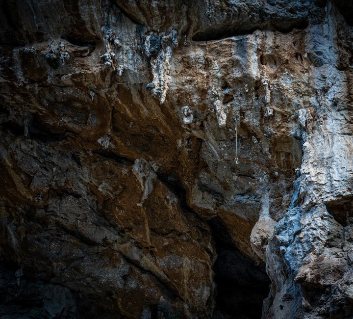 A view of the interior of a limestone cave with stalactites hanging from the ceiling, illuminated by warm, yellowish lighting that highlights the textured, rough surface of the rock formations. The ca